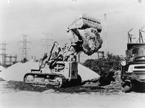 A man is operating the crawler tractor outdoors. The crawler is carrying a large rock towards a truck parked on the right. Power lines are in the background. Caption reads: "International Harvester TD-15 Crawler Tractor with Drott Four-In-One Attachments." Text on back of print reads: "N.M. Pierre, Skokie, Ill."