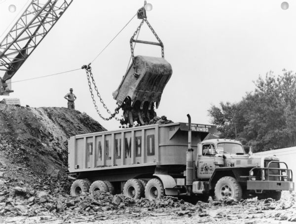 A truck is being loaded from a steam shovel, and a man is standing on the gravel hill behind the truck. Sign on truck reads: "Palumbo." Caption reads: "International Model F-230-D 220 Horsepower Diesel Powered Tractor Truck." Text on back of print reads: "Palumbo Excavating Company, Chicago."
