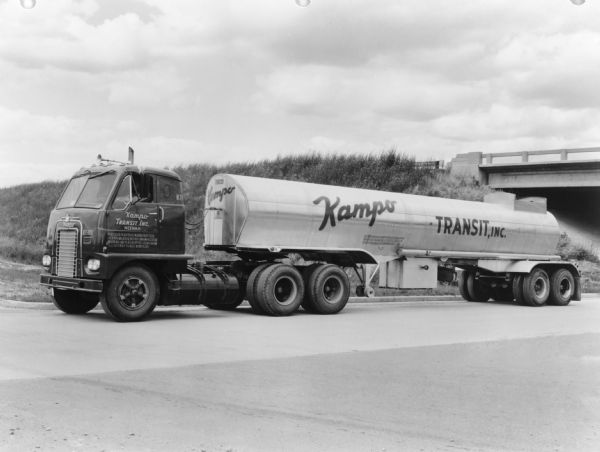View towards driver's side of truck, parked on a road, with a bridge on the far right behind the truck. The sign on the truck reads: "Kampo Transit, Inc. Neenah." Text on back of print reads: "International model DCOT-405 of Kampo Transit Inc."