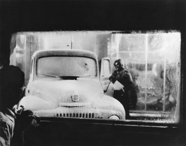 A man is looking through a window towards another man in a cold lab testing a truck. Caption reads: "A parka-clad engineer braves thirty below zero temperature to check an ice-covered truck in the 'cold lab' in International Harvester Company's new motor truck engineering building and laboratories. Bitter cold weather is produced in this laboratory to test cold-weather characteristics of trucks and components. Engineers check starting ability and performance in the cold, and test the starting qualities of fuels and the characteristics of lubricants. Starting motors, cab heaters and defrosters also undergo tests here."
