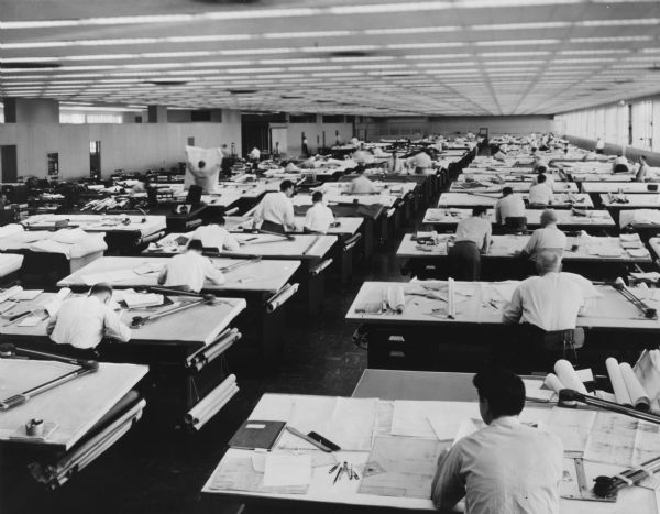 Slightly elevated view from rear of large room looking towards men who are working in a large room sitting in rows of drafting tables.