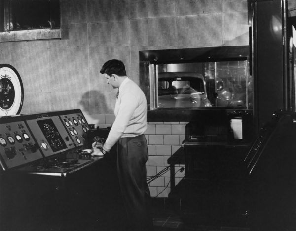 A man is standing at a control board in a room with other equipment. There is a Toledo Scale on the left behind the control board. A window in the wall behind the man looks out to an automobile in another room.