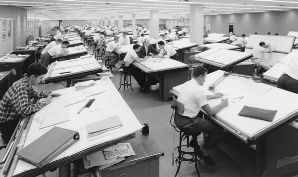 View of large room, with engineers working at drafting tables set up in rows.