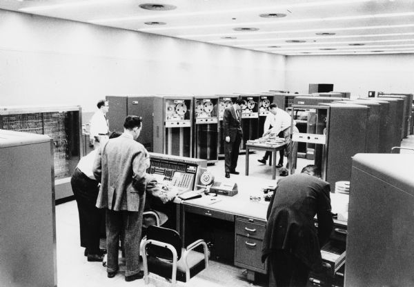 Men are standing in a room with computer equipment. Caption reads: "IBM 705 Electronic Computer. Two of these are available to IH engineering & research personnel."