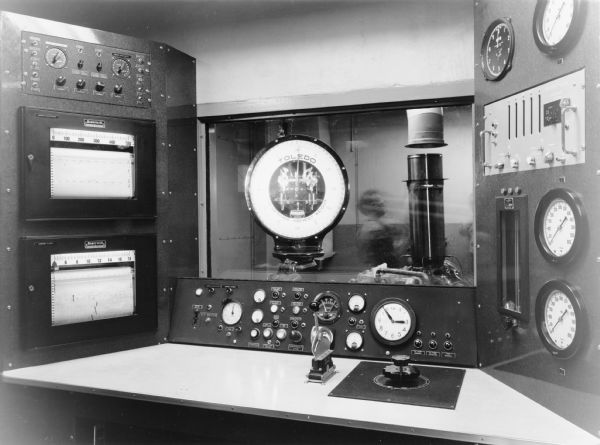Caption reads: "Control Room of Test Cell For Gas Turbine Engines Construction Equipment Eng. Dept." Behind the controls is a window, and set up in the room on the other side is a Toledo Scale. A man (blurred from motion) is also working in the room with machinery.
