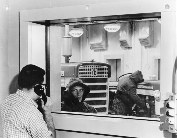 A man in the foreground on the left is holding a telephone receiver while talking with a man who is holding another telephone receiver while standing inside the cold room seen through a window. The man in the cold room is wearing a coat and hood, and another man and two tractors are behind him. Caption reads: "Cold Test Room -50°F (FEREC)."