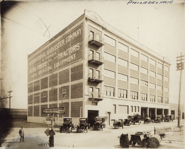 View across street towards the International Harvester's Philadelphia branch house. Railroad tracks are in the background behind the building on the left. A number of automobiles and trucks are parked along the street and sidewalk in front of the building, and a man is standing in the street on the far left. A sign on the side of the building reads: "International Harvester Company of America, Motor Trucks, Tractors, Farm Operating Equipment." Signs in the foreground read, in part: "American Solder & Flux Co., Successors to Hess & Son.," and "Allegheny Chemical Co."