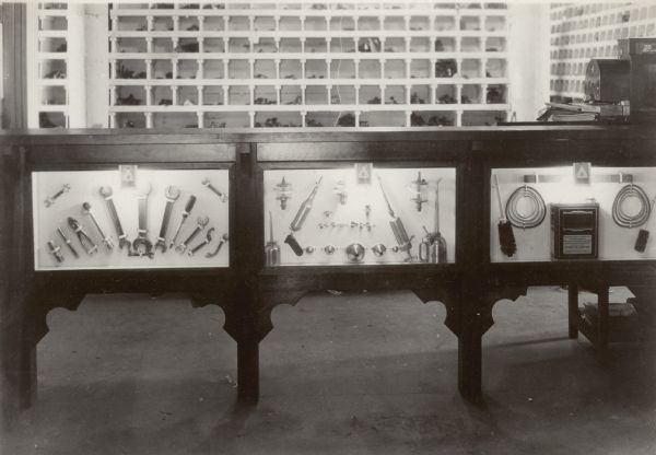 Interior view of display of tools and other products in glass-fronted cases at the service counter. Small signs on each case reads: "IHC Repairs." There is a cash register on the counter on the right. In the background are shelves/cubbies along the walls that hold parts.