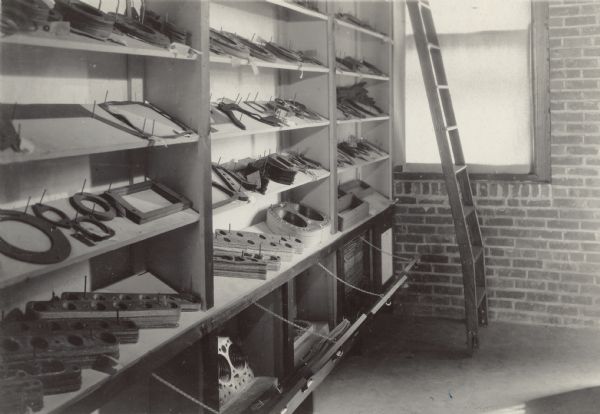 Interior view of display of tools and other products arranged on shelves. The shelves are tilted down to display the parts, which are held in place by pins. In the background in front of a window is a ladder leaning against the shelving system for access to the upper shelves 
