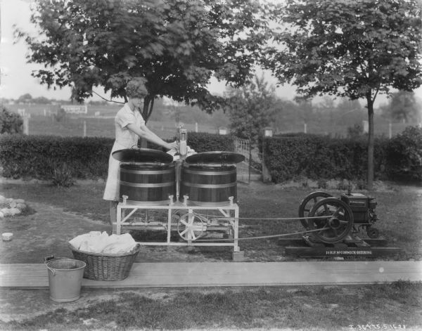 Woman washing clothes outdoors in a yard with a washing machine powered by a McCormick-Deering 1 1/2 H.P. engine.