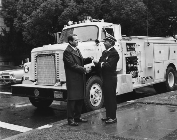 Two men are standing on a sidewalk in front of a fire truck. News release accompanying the photograph is titled: "International Fire Trucks to 10 California Departments".