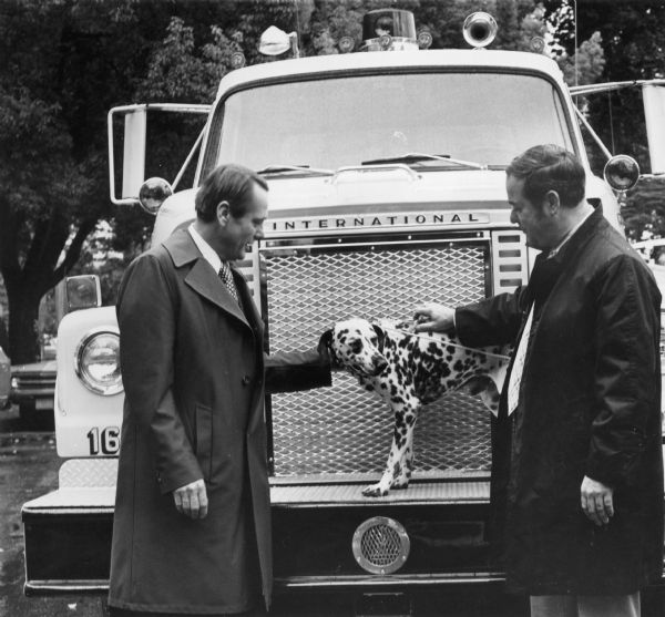 Two men with a Dalmation dog and a fire truck. News release accompanying the photograph reads: "California's Lt. Governor Ed Reinecke (left), recently presented ten International model 2050-A Fleetstar fire trucks to local Chiefs from all over the state. The new units, painted a distinctive lime and yellow color to make them more visible, were equipped with CAT 1160 engines. Also on hand for the presentation was Dick Barrows, Chief, Fire and Rescue Division, Office of Emergency Services."
