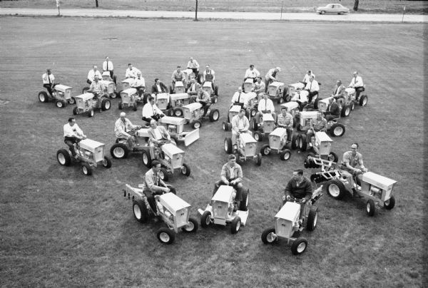 Elevated view of a group of about 32 men sitting on Cub Cadets parked outdoors on a large lawn. An automobile is parked on a road in the background. This is most likely at the Louisville Works factory.