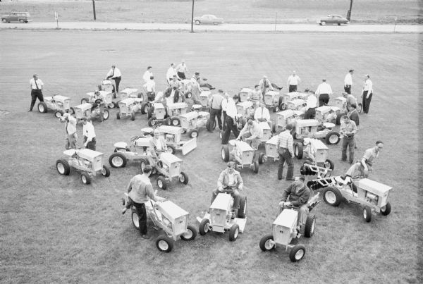 Elevated view of a group men sitting on, getting on, or standing near Cub Cadets parked outdoors on a large lawn. Automobiles are parked on a road in the background. This is most likely at the Louisville Works factory.