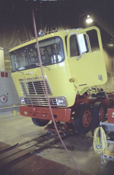 Three-quarter view from front towards left side of cab of truck in the factory. The cab is tipped forward, and the driver's side door is open. A man is working on the chassis behind the cab.