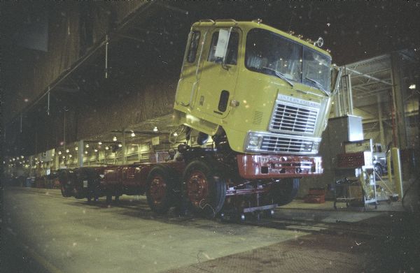 Three-quarter view from front towards right side of cab of truck in the factory. The cab is tipped forward. Two men are working on the chassis behind the cab. Toolboxes are along the wall on the far right.