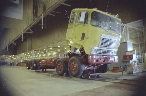 Three-quarter view from front towards right side of cab of truck in the factory. The cab is tipped forward. Two men are working on the chassis behind the cab. Toolboxes are along the wall on the far right.