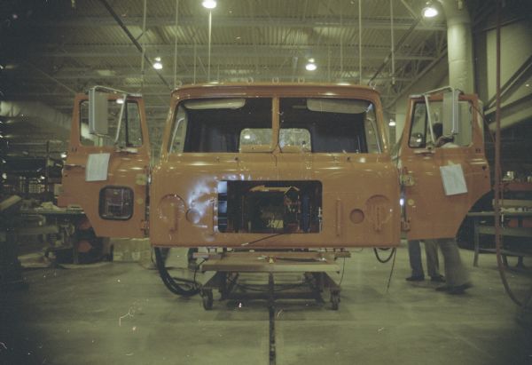 View towards the front of a truck cab body in the factory which is sitting on a stand. Two man are standing on the right behind the open driver's side door. The passenger side door is also open. Another man is working behind the cab body which has an open section on the front.