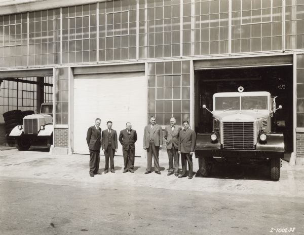 Outdoor group portrait of eight men standing outside of the Emeryville factory with a truck parked in the open doorway. Another truck is parked in an open doorway on the left. Caption reads: "The first four heavy-duty 'Western' models produced at International Harvester's Emeryville, Calif., Works were these 215-inch wheelbase model W-4064-H unites built for International Cementers, Inc., of Long Beach, California. Oil well cementing equipment will be mounted on these units. Individuals in the picture are: H. E. Straub, General Foremen; K. A. Roush, Foreman of Cab Foundry and pattern Department; Merrill Bell, Foreman, Machine Shop; C. W. Gallogley, Foreman, Frame Shop; A. W. Engstrom, Works Manager; and A. S. Busselle, Parts Manager. All of Emeryville Works."