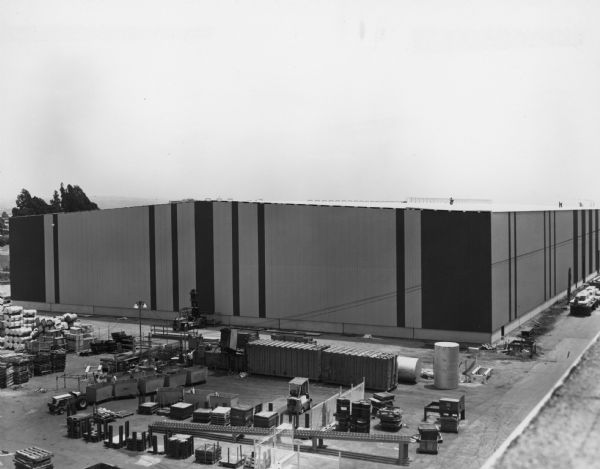 Elevated view of a new warehouse. Men are working on the ground and on the roof. Caption on back reads: "1973-1974. Progress Report".