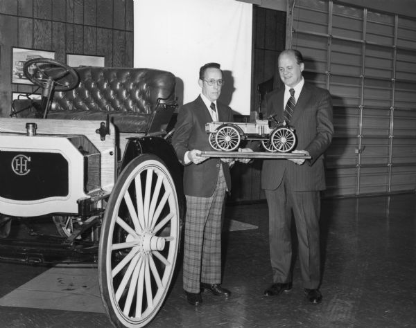 Attached letter from the Springfield Plant in Ohio reads, in part: "Attached are photos of Robert E. Brown, the model builder, presenting the model to Mr. Mazurek for G. O. Archives. The model will be carefully packed and covered with a plexiglas display cover before it is sent to Archives." Next to them on the left is an auto wagon. A large, roll-up garage door that is closed is behind them on the right.
