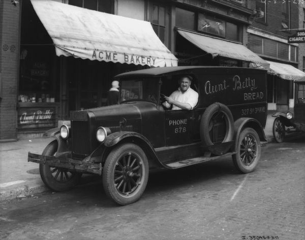 A man is sitting in the driver's seat of a delivery truck parked at the curb in front of Acme Bakery. The sign painted on the truck reads: "Aunt Betty Bread."