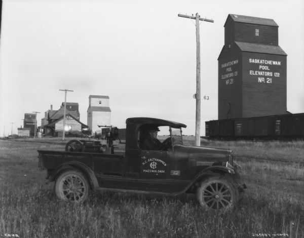 A man is sitting in a truck with a cream separator in the truck bed. Sign on the truck reads: "H.C. Salthammer, Mazenod. Sask. IHC." In the background are grain elevators and railroad cars. The sign on the grain elevator reads: "Saskatchewan Pool Elevator Ltd. No. 21."