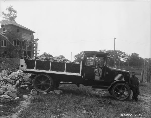 A man is standing at the front of a construction truck with his thumbs hooked in the beltloops of his pants. There are large rocks in the bed of the truck. The sign painted on the side of the truck reads: "James Piro & Sons." In the background is scaffolding around a building.