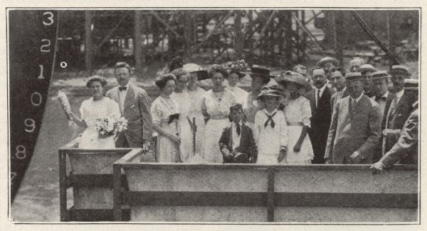 A group portrait of the formally attired launching party about to christen the ship S.S. <i>Harvester</i> on the day she was launched. The woman on the left holds a champagne bottle at the ready. 