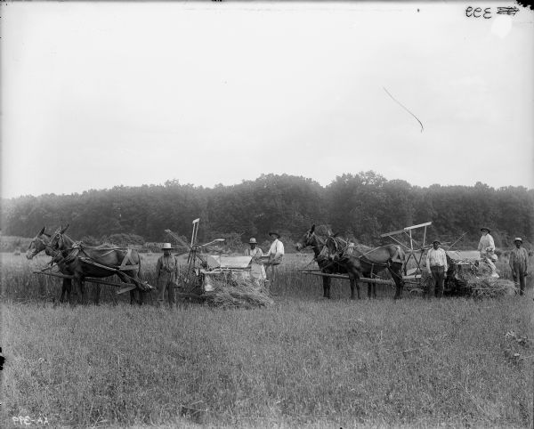 Six men in a field with two mule-drawn binders. Two of the men are sitting on the binders. Trees are in the background.