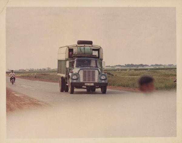 International Harvester truck on a road in Vietnam. A man is riding a motor scooter behind the truck on the left. In the background is a field, and beyond a village. The trucks were used to haul goods from the different provinces to Saigon, a city of three million people, many of whom have fled their country homes to the city because of the war.  Report states: "Trucks used to carry vegetables from Da Lat (Central Highlands) to Saigon, over a distance of 300 kilometers. To travel from Da Lat to Saigon takes 6.5-7 hours. The roads are in good-fair condition." Description of equipment: City: Cholon. Loadstar 1700, 3 years old, standard; RA-146 Used."