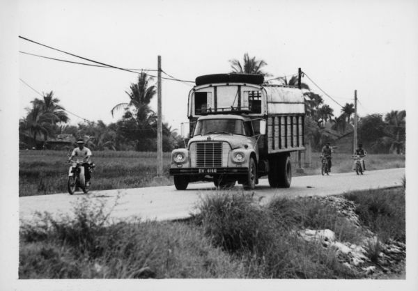 View from side of road towards a man driving an International Harvester truck on a road in Vietnam. A man riding a scooter is in front of the truck, and two more people riding scooters are behind the truck on the right. The trucks were used to haul goods from the different provinces to Saigon, a city of three million people, many of whom have fled their country homes to the city because of the war. Report states: "Trucks used to carry vegetables from Da Lat (Central Highlands) to Saigon, over a distance of 300 kilometers. To travel from Da Lat to Saigon takes 6.5-7 hours. The roads are in good-fair condition." Description of equipment: City: Saigon. Loadstar 1700, 3 years old.