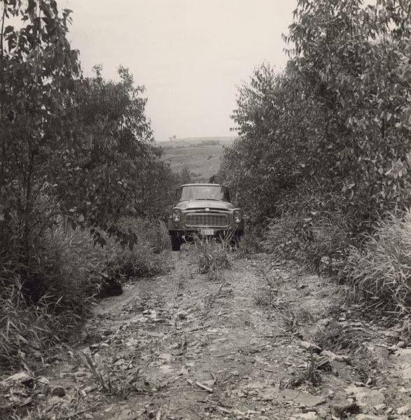 View towards the front of the truck moving uphill through dense vegetation. In the distance are hills. Caption reads: "Travelette of the Malaria Eradication Service maneuvering a difficult road into living quarters for workmen on a large sugar plantation in the region of Piracicaba, state of São Paulo.