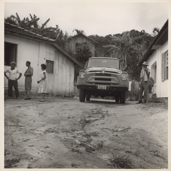 Truck parked among buildings, with people standing nearby. Caption reads: "Malaria eradication personnel talking to farmer in the coastal regions north of Santos before starting to spray his home and other buildings with the DDT solution which will [sic] the mosquitos, not only at spraying time but for a long time afterwards."