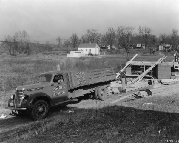 A man is standing and unloaded lumber near the back of an International D-35 truck owned by Campbell Coal. The driver's side door has the text: "Campbell Coal Co." The side of the bed of the truck has text that reads: "Millwork Lumber Building Supplies." Behind the truck on the right two men iare standing on top of a partially constructed brick building.