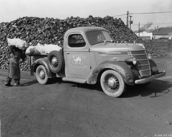 A man (face obscured) carrying a sack of coal on his shoulder is standing near the back of the bed of an International D-2 truck owned by Campbell Coal. The passenger side door features an illustration of a camel and the text: "Campbell Coal 'Most Heat Per Dollar.'" A large pile of coal is behind the truck near a fence. In the distance on the right are industrial buildings.