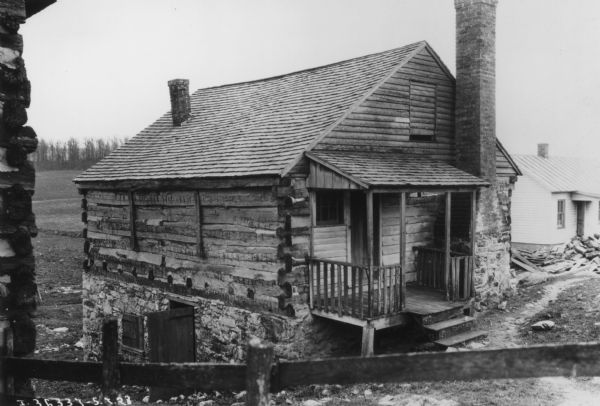 View over wood fence looking down towards the front and left side of a log building with a peaked roof, second story, and a stone foundation built into the side of a hill. At the front is a small porch with a door and a window, and an exterior chimney at ground level next to the porch. Another chimney extends through the roof near the back of the building on the left. At lower left below the hill is the stone foundation, window with a hinged wood cover, and an open wood door in the wall. The corner of a log building is in the left foreground. Another building is in the background on the right. Piles of wood and lumber are laying in the yard between the two buildings. In the background are fields with trees along the edge.