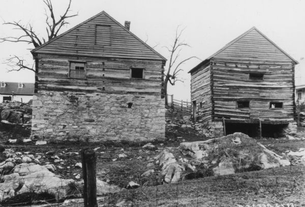 View across fence looking up toward the backs of two buildings built into a hill. The Blacksmith Shop on the left has a stone foundation, two windows on the first floor, and a wood covered window near the top of the peaked roof. The Mill Building on the right has a peaked roof, a small stone foundation with openings at the bottom, three small, horizontal openings on the first floor, and a door on the left side wall. A building is in the background on the far right, and a barn is on the hill in the background on the left.