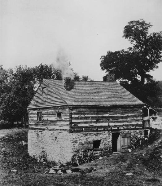 View down hill looking towards the side and back of a log building with stone foundation built into the side of a hill. Smoke is rising from the chimney extending from behind the peaked roof near the front of the building. There is another chimney in behind the peaked roof. Wagon wheels rest against the side of the foundation, and there is an open window and door in the foundation. The railing of a small porch on the first floor is at the front of the building on the right.