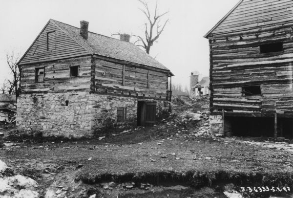View towards the backs of two log buildings built into the side of a hill. The building on the left has a stone foundation, two windows on the first floor at the back, and a wood covered window near the top of the peaked roof. There is an open door and a window in the stone foundation, and at the front right corner is part of a railing and a roof over the porch which is at the front of the building. On the right, the other log building, partially seen on the right foreground, has a peaked roof, a small stone foundation with openings at the bottom, and two small, horizontal openings on the first floor. Between the buildings in the background is a man carrying something on his way towards a barn and a silo. Other men are working in the barnyard.