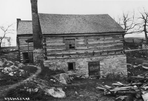 View looking downhill toward the side of a log building with stone foundation built into the side of a hill at Walnut Grove. A path is on the left leading to the front of the building, which has an exterior chimney and a small, roofed porch with railings. There is a window on the first floor, and a door and window below in the foundation wall. Lumber and wood is piled in the yard on the right. The corner of another log building can be seen in the background on left just behind the building in the foreground, with a peaked roof and an exterior chimney. In the background are fields and trees.