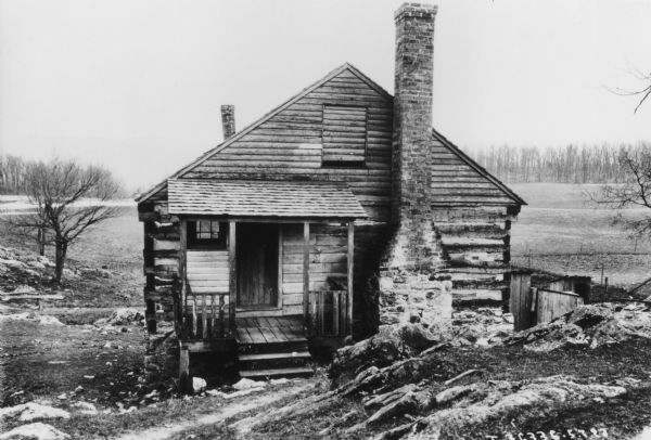 View looking down towards the front of a log building built into the side of a hill. A small roofed porch with a window and door is on the left, and an exterior chimney is next to the porch just right of the peaked roof. A wood covered window is centered near the peaked roof. Another chimney extends from the back of the roof on the left. In the background are fields and trees.