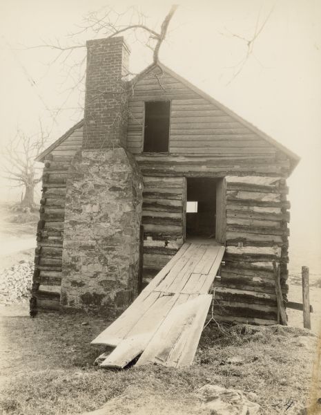 View of the front of the McCormick Mill at Walnut Grove, with a wooden walkway up to the open door or window on the right. An exterior chimney is to the left of the entrance, and an open window is above near the peaked roof. The mill is built into the side of a hill. Trees are below in the background. Accompanying note reads: "Original McCormick Mill Building, constructed about 1800, as it appeared in the summer of 1937. Note the leaning chimney which is forcing the building off its foundation."