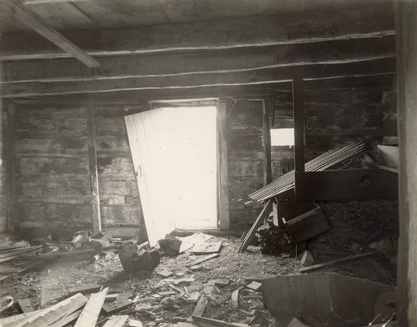 Interior view of building, looking towards open door and small window. Debris is on the floor, and the back corner on the right has broken shelves and a stack of wood below.