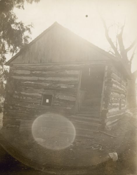 Exterior view of front and right side of log building. Wood steps lead up to the open doorway on the right, and the steps continue inside up to the floor level. A small window is in the center. The building has a peaked roof. On the right side of the building is a window into the lower level.