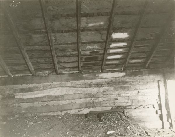Interior view towards side of log building. In the right corner are steps leading down to the right towards a door. Beams are supporting the wood floor above.