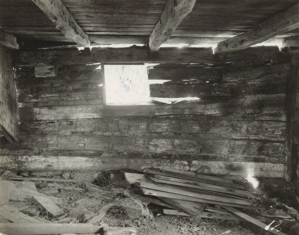 Interior view of log building. A window opening is just below the ceiling. Debris is on the floor. There are openings between the logs to the outdoors.