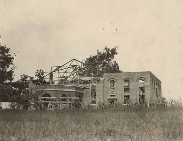 View across field towards men working on a brick building under construction. Scaffolding is set up around the building, and frames for the roof are exposed.