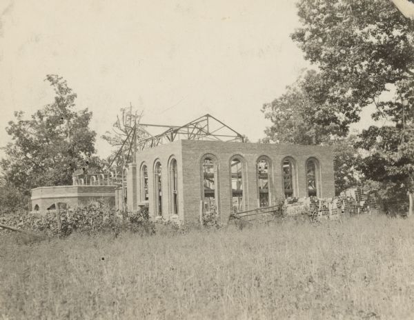 View across field towards a brick building under construction. Frames for the roof are exposed. A man is working inside one of the arched window frames.