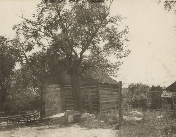 View towards the mill. There is an exterior chimney to the left of the door, and a wooden walkway leads up to the closed door on the right. A fence leads to the building from the right, and another building is behind the fence. In the background on the left are wood railings along a road or bridge.